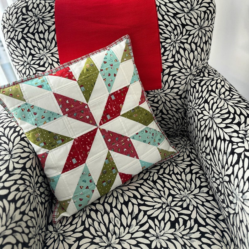 May include: A decorative pillow with a starburst pattern in red, green, and blue geometric shapes. The pillow is on a chair with a black and white floral pattern, and a red throw is draped over the back.