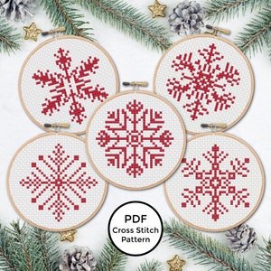 Puede incluir: Cinco patrones de punto de cruz de copo de nieve rojo en aros de bordado. Los patrones están sobre un fondo blanco y los aros están sobre una superficie blanca. El texto "PDF Cross Stitch Pattern" está debajo de los aros.