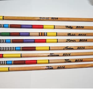 May include: Seven wooden arrows with stone arrowheads. Each arrow has a different color scheme, including blue, yellow, red, and purple bands. Names and the year 2016 are written on each arrow.