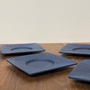 May include: Four blue square ceramic coasters with a raised centre circle. The coasters are on a wooden surface.