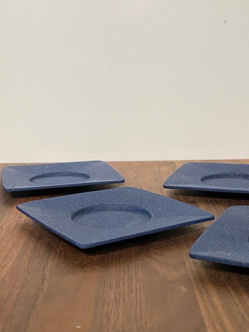 May include: Four blue square ceramic coasters with a raised centre circle. The coasters are on a wooden surface.