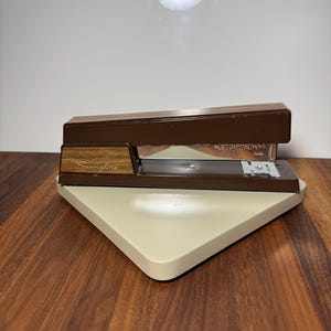 May include: A brown and beige Swingline stapler on a beige base. The stapler has a wood grain accent and the text "Swingline" is visible. The base is a light beige color and is on a wooden surface.