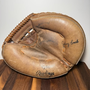 Peut inclure: Un gant de receveur de baseball Rawlings en cuir marron vintage portant les inscriptions Professional Model, Deep Well Pocket, Hinged Pad, CJ-50 et la signature Johnny Bench gravée sur la surface avec un laçage visible sur le bord extérieur.