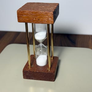 May include: An hourglass featuring a clear glass bulb filled with white sand, positioned between two square wooden bases. Four thin, polished gold-tone metal rods connect the top and bottom wooden pieces, creating a vertical frame.