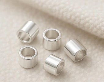 May include: Five small, cylindrical silver beads with a smooth, polished finish. Each bead has a hole running through the center, and they are scattered on a soft, off-white surface. These beads are ideal for jewelry making.