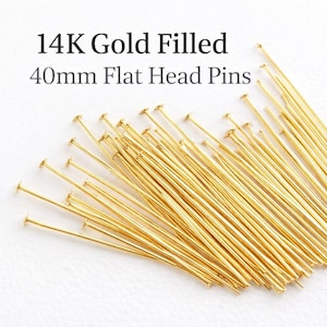 Puede incluir: Un conjunto de alfileres dorados de cabeza plana, cada uno de aproximadamente 40 mm de largo, dispuestos sobre una superficie blanca. El texto negro indica "14K Gold Filled". Estos alfileres se utilizan probablemente para la fabricación de joyas o manualidades.