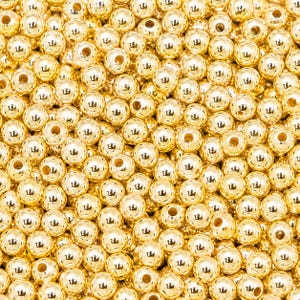 Puede incluir: Primer plano de numerosas cuentas brillantes de color dorado. Cada cuenta es redonda con un pequeño agujero que atraviesa el centro. Las cuentas están densamente empaquetadas, creando una superficie texturizada y reflectante. Estas cuentas son adecuadas para la fabricación de joyas y manualidades.