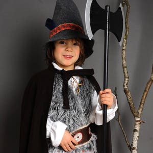 May include: A costume consisting of a dark gray felt hat with a patterned band, a black cape, a white long-sleeved shirt, a gray faux fur vest, brown trousers, and beige leg warmers with red ties. A black double-headed toy axe is held in hand.