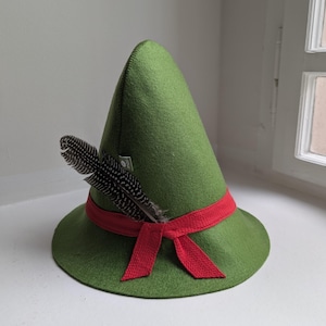 May include: A pointed green felt hat featuring a red fabric band tied in a bow at the front. A single black and white speckled feather is tucked into the red band on the left side. A small white rectangular brand tag is visible on the side.