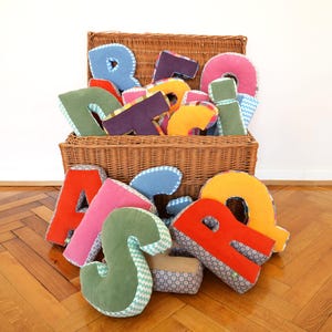 May include: A collection of plush fabric letters in various colours and patterns, including red, orange, pink, blue, and green, overflowing from a large brown woven wicker basket. The soft, stuffed alphabet characters feature contrasting side panels.