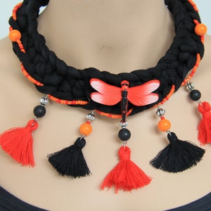 Può includere: Collana choker in tessuto nero con perline arancioni e nere, un ciondolo libellula rosso e nappe nere e rosse.