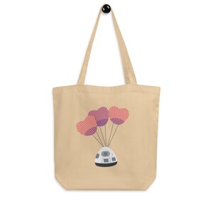 May include: A beige tote bag with a white space capsule design. The capsule is being lifted by three purple and red striped heart-shaped balloons.