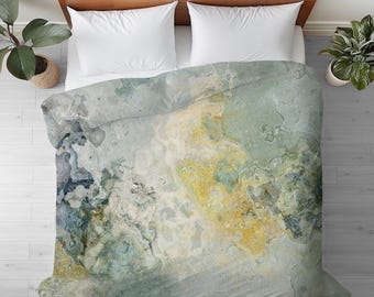 Può includere: Un letto con testiera in legno e cuscini bianchi. Il letto è coperto da un copripiumino con un design astratto nei toni del blu, giallo e verde, che ricorda un motivo marmorizzato. Piante in vaso sono su entrambi i lati del letto.