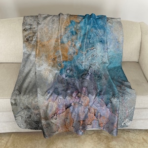 May include: A soft, plush throw blanket with an abstract design featuring shades of gray, blue, and brown. The design resembles a marbled or textured surface.