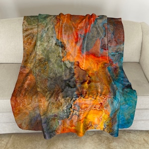 May include: Abstract throw blanket with a colorful design featuring shades of orange, yellow, blue, and gray. The blanket has a soft, plush texture.