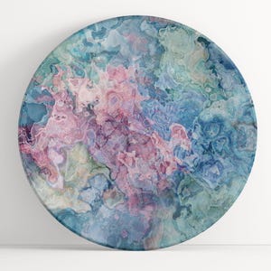 May include: Round decorative plate with an abstract design. The plate features a swirling pattern of pink, blue, and green hues, creating a marbled effect. The plate is set against a white background.