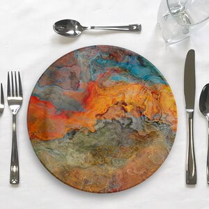 Puede incluir: Un plato de cena redondo con un dise&ntilde;o abstracto colorido que presenta remolinos de naranja, azul, gris y marr&oacute;n. El plato est&aacute; colocado sobre un mantel blanco con cubiertos.
