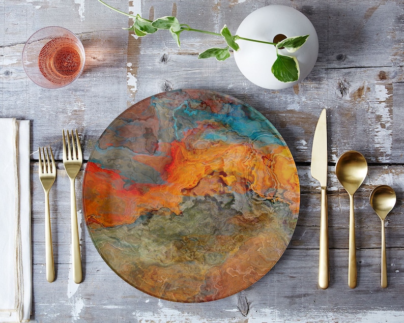 Puede incluir: Un plato redondo y colorido con un dise&ntilde;o de m&aacute;rmol en tonos naranja, marr&oacute;n, azul y verde. El plato est&aacute; colocado sobre una mesa de madera con cubiertos de oro y una copa de vino.