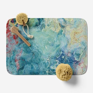 May include: A blue, green, and yellow abstract bath mat with a marbled design. The mat is on a white surface with two bath sponges and a wooden bath brush.