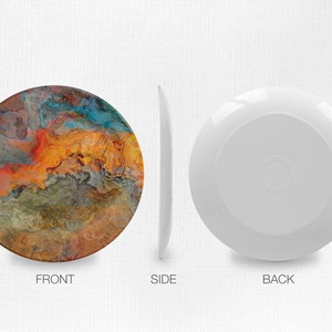 Puede incluir: Un plato redondo de cer&aacute;mica con un dise&ntilde;o abstracto colorido que presenta tonos de naranja, amarillo, azul y marr&oacute;n. El plato es blanco en la parte posterior.