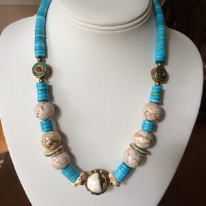May include: A turquoise and white beaded necklace with a large white stone pendant. The necklace features small, round, turquoise beads and larger, round, white beads with brown and red markings. The pendant is a large, round, white stone with brown and red markings.