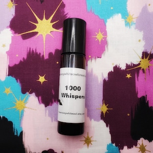 May include: A small, clear glass bottle with a black roll-on top. The bottle is labelled "1000 Whispers" and has a white background. The bottle is sitting on a colourful fabric with a star pattern.