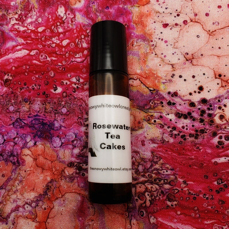 May include: A brown glass roller bottle with a white label that reads "Rosewater Tea Cakes" and a website address. The bottle is sitting on a pink and white abstract background.