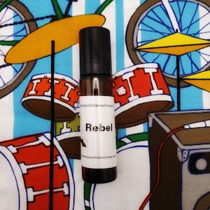 May include: A brown glass roll-on bottle with a black cap and a white label that says "Rebel". The bottle is sitting on a colorful fabric background with a drum set pattern.