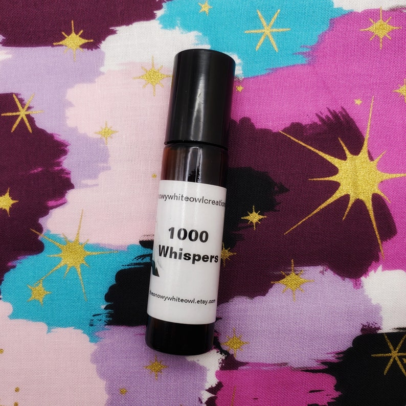 May include: A small glass bottle with a black roll-on top. The bottle is labelled "1000 Whispers" and has a white background with black text. The bottle is sitting on a colourful fabric with a star pattern.