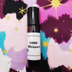 May include: A small, clear glass bottle with a black roll-on top. The bottle is labelled "1000 Whispers" and has a white background with black text. The bottle is sitting on a colourful fabric with a star pattern.