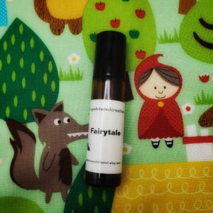 May include: A small, clear glass bottle with a black roll-on top. The bottle is filled with a dark liquid and has a white label with the text "Fairytale" on it. The bottle is sitting on a green and yellow fabric with a cartoon design.