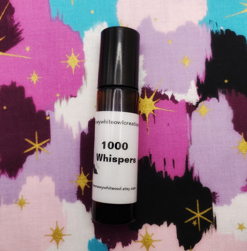 May include: A small brown glass bottle with a black rollerball top. The bottle has a white label with the text "1000 Whispers" and the website address "snowywhiteowl.etsy.com".