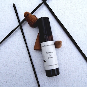 May include: A black essential oil roller bottle labeled "Incense in Snow" with a white label, surrounded by incense sticks and cones. The background is a textured white surface. The bottle is approximately 3 inches tall.