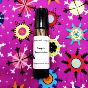 May include: A small, brown glass roll-on bottle with a black cap, labeled "Purple Persuasion." The bottle sits on a vibrant purple fabric patterned with colorful starburst designs. The text "snowywhiteowl.etsy.com" is also visible.