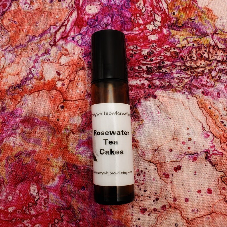 May include: A glass bottle with a white label that reads "Rosewater Tea Cakes" and a website address. The bottle is on a pink and white marbled background.