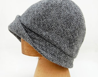 May include: A gray tweed cloche hat with a wide brim and a slightly slouched crown. The hat is displayed on a wooden hat form.