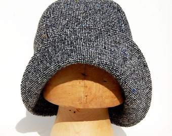 May include: A black and white tweed cloche hat with a wide brim. The hat is sitting on a wooden hat form.