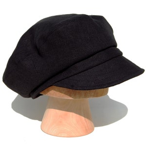 May include: A black linen newsboy cap with a short, stiff peak. The cap is displayed on a wooden hat stand against a white background. The hat has a rounded crown and a structured, slightly angled peak.