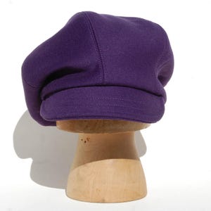 May include: A purple beret-style hat with a rounded crown and a structured brim. The hat is made of a soft, felt-like material and is displayed on a wooden hat stand. The hat has a classic design and is suitable for various occasions.