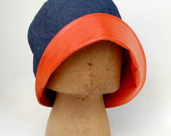 Könnte beinhalten: Ein Cloche-Hut aus Jeans mit einem leuchtend orangefarbenen Besatz. Der Hut befindet sich auf einer Hutform aus Holz.