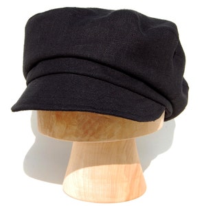 May include: A black newsboy cap, featuring a rounded crown and a small, stiff peak. The cap is made from a textured material and is displayed on a wooden hat stand. The dark color of the hat contrasts with the bright background.