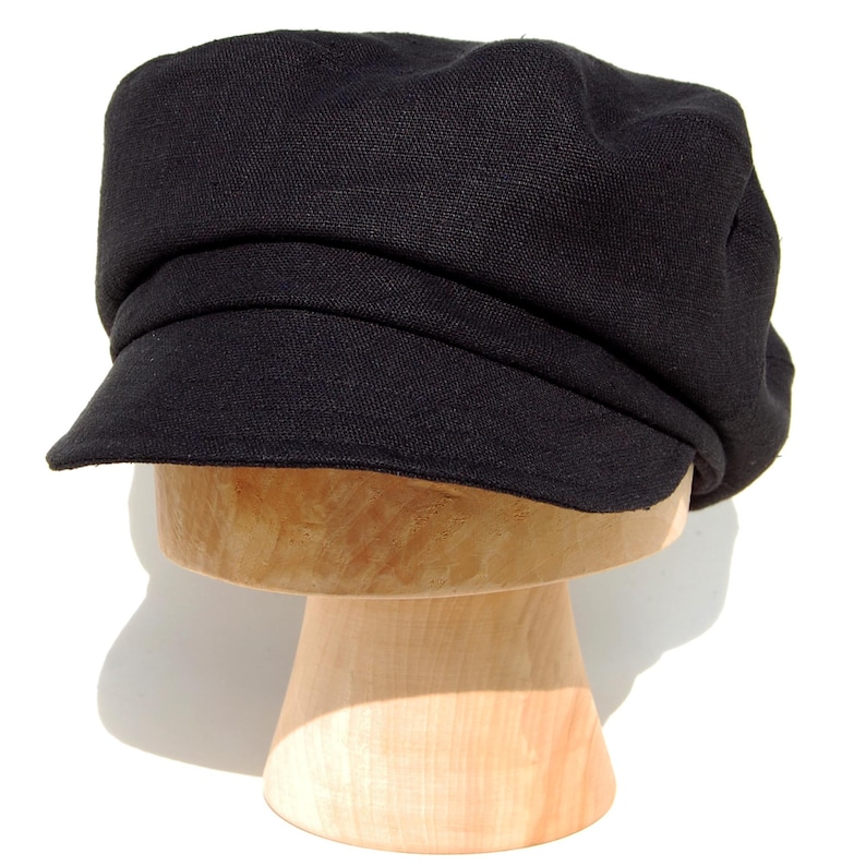 May include: A black newsboy cap, featuring a rounded crown and a small, stiff peak. The cap is made from a textured material and is displayed on a wooden hat stand. The dark color of the hat contrasts with the bright background.