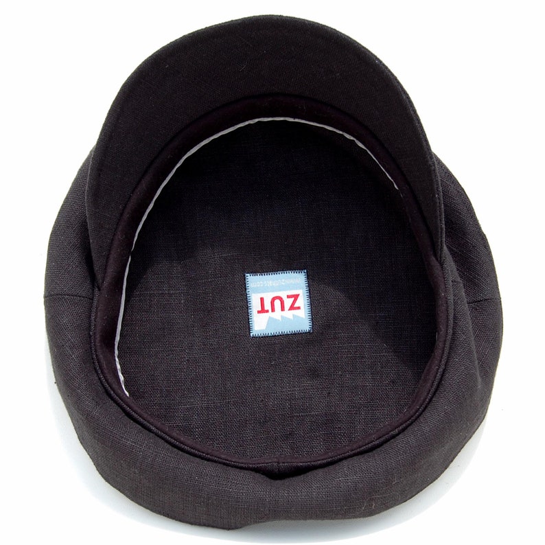 May include: A dark brown newsboy cap with a rounded crown and a short, stiff peak. The interior has a white band and a small label with the text "ZUT" in red letters on a blue and white background. The cap is made of a textured fabric.