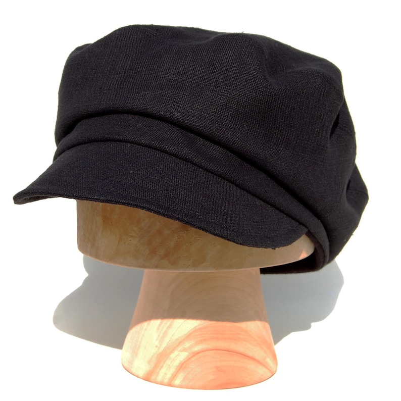 May include: A black newsboy cap with a short peak, presented on a wooden hat block. The cap features a rounded crown and a structured design. The hat is made of a textured fabric.