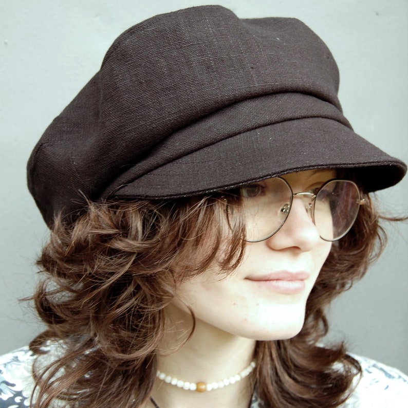May include: A dark brown newsboy cap with a rounded crown and a small, stiff brim. The cap is made of a textured fabric and is worn at an angle. The person is wearing round, gold-rimmed glasses and a pearl necklace.
