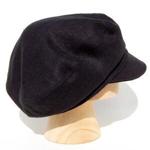 May include: A black linen newsboy cap with a short peak. The cap is displayed on a wooden hat stand against a white background. The hat has a rounded crown and a structured peak.
