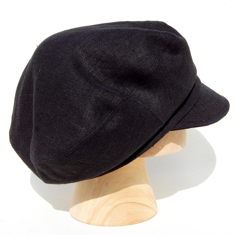 May include: A black linen newsboy cap with a short peak. The cap is displayed on a wooden hat stand against a white background. The hat has a rounded crown and a structured peak.