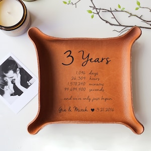 May include: A square, tan leather tray with rounded corners. The tray has the text "3 Years" and a list of time measurements. Below the list is the text "...and we've only just begun" and the names "Gia & Mitch" with a date.