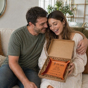 May include: A couple smiles while looking at a gift box. Inside, a brown leather tray is visible, inscribed with the words "3 YEARS" and a list of dates and names. The tray is nestled in a cardboard box with shredded paper.