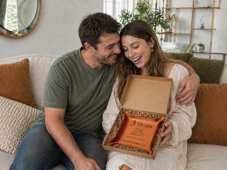 May include: A couple smiles while looking at a gift box. Inside, a brown leather tray is visible, inscribed with the words "3 YEARS" and a list of dates and names. The tray is nestled in a cardboard box with shredded paper.